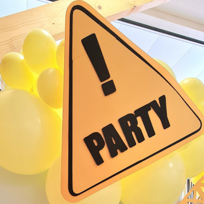 Sinal amarelo triangular com texto '! PARTY' rodeado por balões amarelos