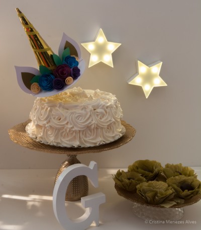 Bolo branco decorado com creme, chifre dourado e flores de papel, estrelas luminosas e letra decorativa G