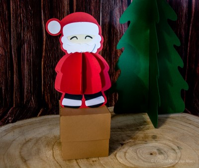 Figura de Pai Natal vermelho e branco em cartão 3D sobre caixa castanha com árvore verde cartesiana e fundo madeira