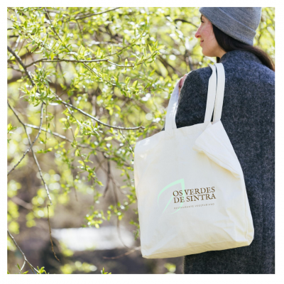 Bolsa de tecido creme com texto OS VERDES DE SINTRA que uma pessoa com casaco cinzento usa ao ombro num ambiente exterior com árvores