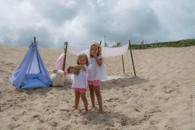Duas meninas em roupa clara na areia junto a tendas de tecido azul e branco com almofadas e urso de pelúcia