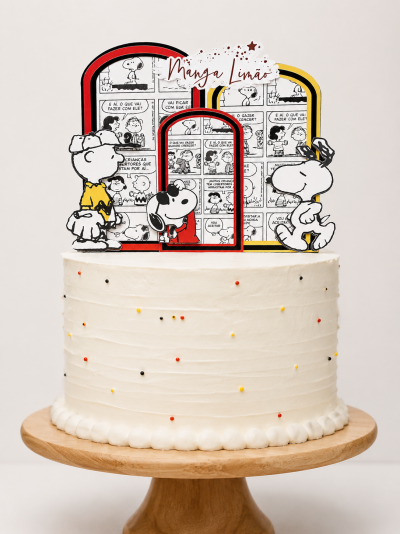 Bolo branco decorado com confeitos coloridos e decoração de topo com personagens Snoopy e Charlie Brown.