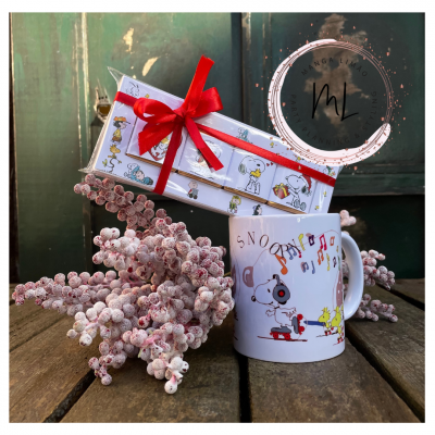 Caneca branca com Snoopy e caixa com laço vermelho sobre mesa de madeira e flores esbranquiçadas