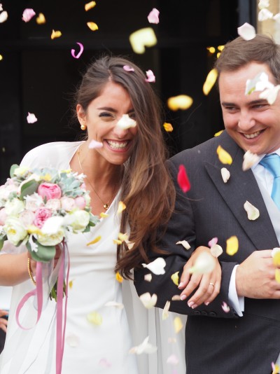 Noiva com vestido branco e ramo de flores e noivo de fato cinzento sorrindo com pétalas caindo