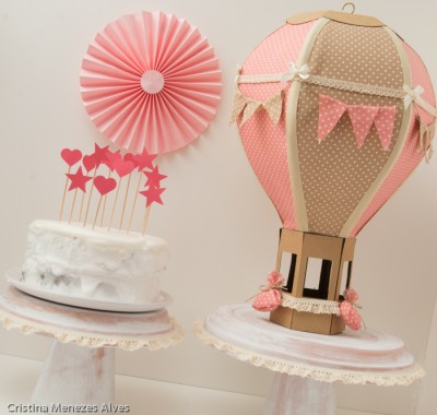 Bolos decorados em suportes brancos com detalhes rendados, um com toppers cor de rosa e outro com balão de ar quente decorativo em papel.