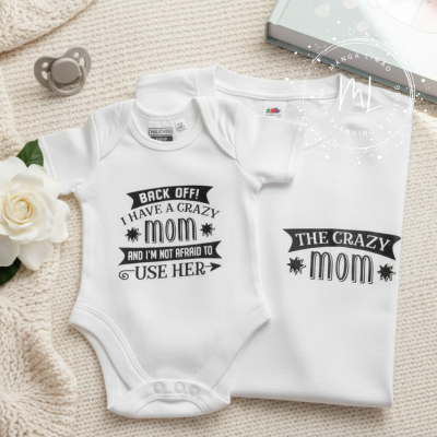 Conjunto de dois bodies de bebé brancos com texto preto sobre superfície bege com chupeta cinzenta e flor branca ao lado.