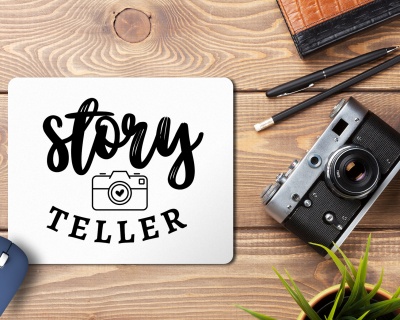 Mousepad com texto story TELLER e câmara fotográfica, sobre mesa de madeira com câmara analógica, canetas, caderno e planta.