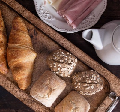 Tabuleiro de madeira com pães variados, prato com fiambre e jarro branco de cerâmica