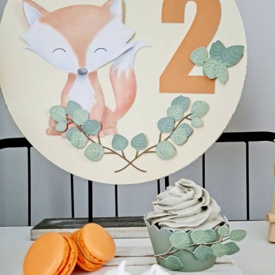 Placa decorativa com raposa, número 2 e ramos verdes, macaron laranja e cupcake cinzento em superfície branca