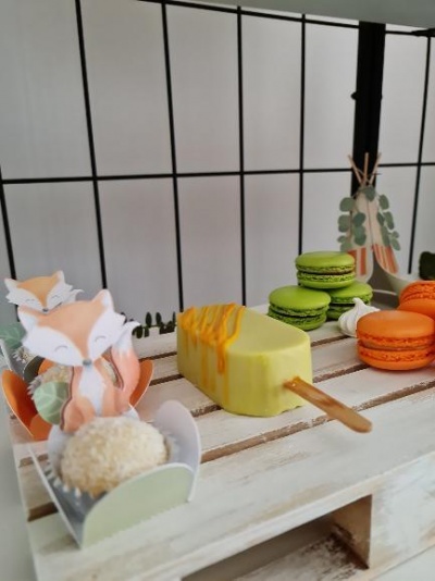 Doces variados sobre prateleira branca, incluindo picolé amarelo, macarons e embalagem decorativa com raposas.