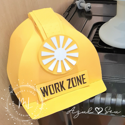 Capacete amarelo com etiqueta 'WORK ZONE' numa área próxima a uma superfície preta e parede de madeira