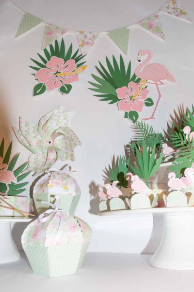 Decoração de festa tropical com flamingos cor-de-rosa, flores, folhas e elementos em papel com padrão floral em fundo branco.