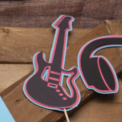 Figura de guitarra de papel preta com detalhes vermelhos e azuis em palito sobre madeira rústica