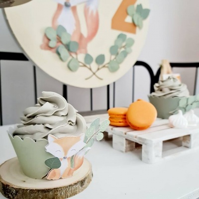 Duas bolos muffin com decoração de raposas e folhas sobre mesa branca com macarons laranja