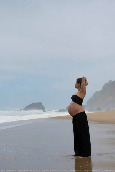 Mulher grávida de top e saia pretos na praia com mar e pedras ao fundo