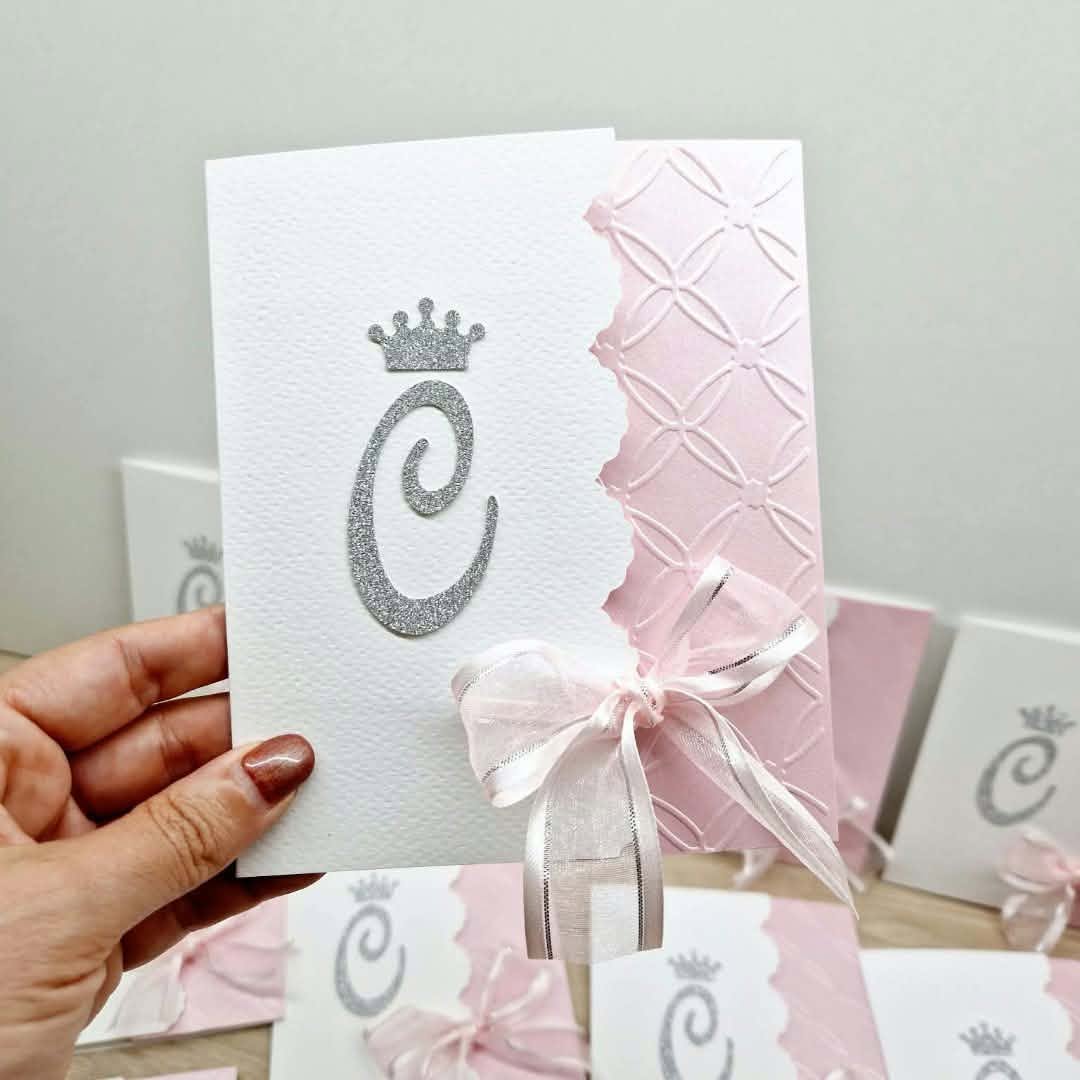 Convite de Comunhão Rosa Perolado em Relevo com Coroa Convite branco e rosa com laço branco e inicial C em glitter prateado, fundo com outros convites similares