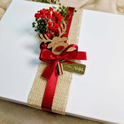 Caixa branca decorada com laço vermelho, fita bege, flores secas e ornamento de rena com etiqueta FELIZ NATAL