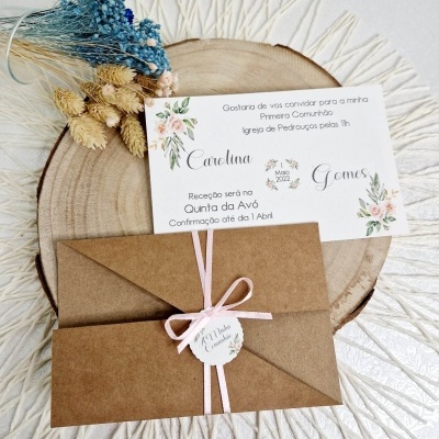 Convite para Primeira Comunhão com envelope castanho, fita rosa e flores secas em fundo branco texturizado.