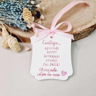 Etiqueta de papel branca com texto em rosa pendurada por fita rosa sobre madeira e flores secas