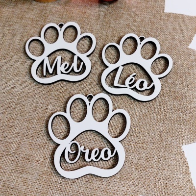 Placas decorativas em forma de pata de animal com nomes Mel, Léo e Oreo sobre tecido bege.