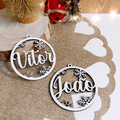 Ornamentos de Natal em madeira branca com os nomes Vitor e João e flocos de neve, sobre toalha de juta e bordado branco.