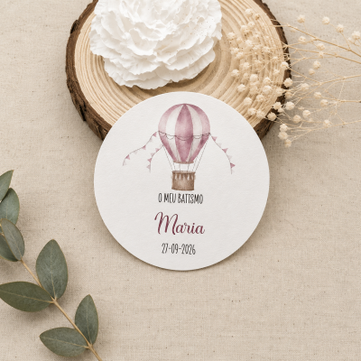 Convite redondo para batismo com balão de ar quente e texto em papel branco sobre tecido bege.
