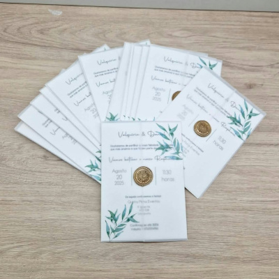 Convites de casamento em papel branco com detalhes verdes e selo dourado sobre mesa de madeira.