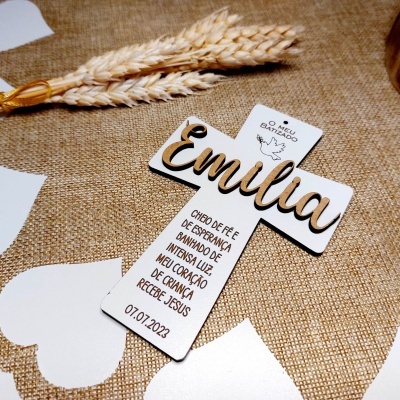 Cruz decorativa branca com nome Emília e texto impresso sobre tecido castanho com corações brancos e espigas de trigo
