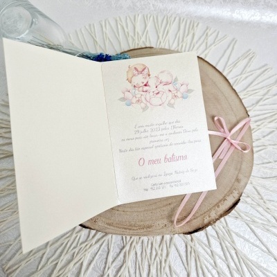 Cartão de batismo com ilustração de bebés e texto colocado sobre base de madeira e fita cor-de-rosa