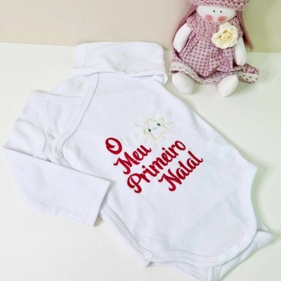 Body branco para bebé com inscrição 'O Meu Primeiro Natal' e boneca de pano cor-de-rosa ao lado.