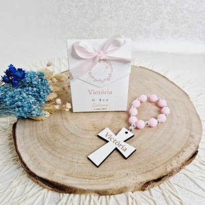 Pulseira com cruz branca em madeira e contas rosa sobre tronco de árvore com cartão de batismo e flores secas
