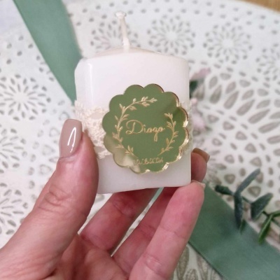 Vela branca pequena com etiqueta verde escura e laço bege sendo segurada por uma mão