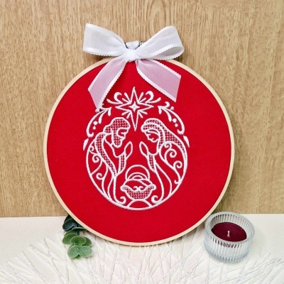 Aro de bordado vermelho com presépio branco e laço branco junto a vela e folhas verdes.