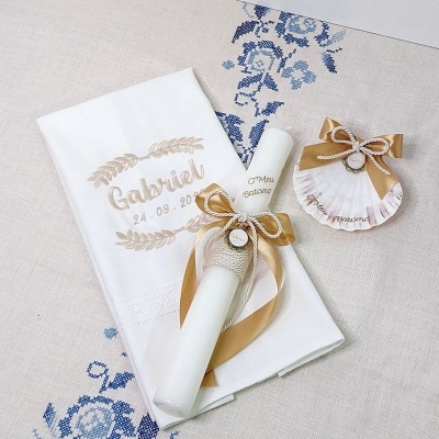 Conjunto de vela, toalha e concha decorativa bordados com nome Gabriel e data, em tons branco, bege e dourado num fundo com padrão azul.