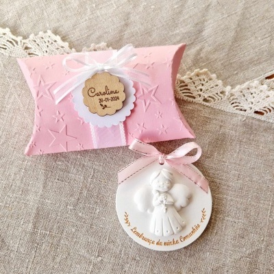 Embalagem rosa com textura de estrelas, etiqueta com nome e data, e medalhão branco com anjo e fita rosa sobre tecido bege