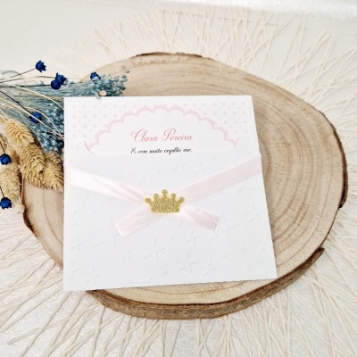 Cartão branco com textura de estrelas, fita rosa e coroa dourada sobre madeira clara e raminhos secos.
