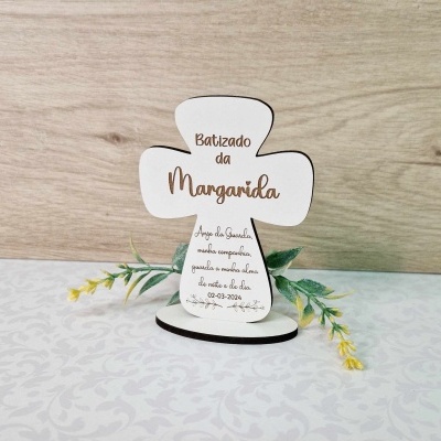 Cruz decorativa branca com texto para batizado, em suporte redondo, sobre superfície branca com padrão e fundo madeira clara