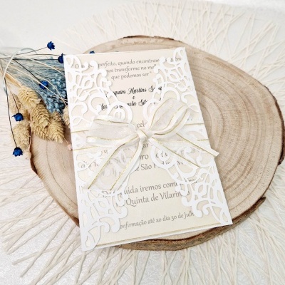 Convite elegante em papel branco cortado a laser com laço de cetim, sobre disco de madeira natural com flores secas azuis e amarelas.