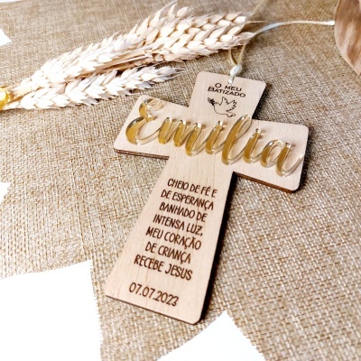 Cruz de madeira clara com nome Emília em letras douradas sobre tecido bege com espigas de trigo