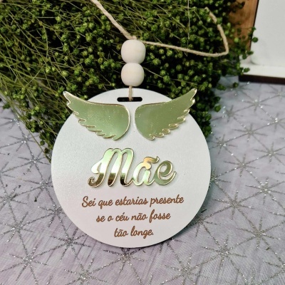 Ornamento redondo branco com asas verdes e texto em dourado e castanho pendurado em cordão com contas de madeira