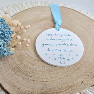 Placa redonda branca com fita azul decorativa e texto em azul apoiada numa base de madeira com flores secas