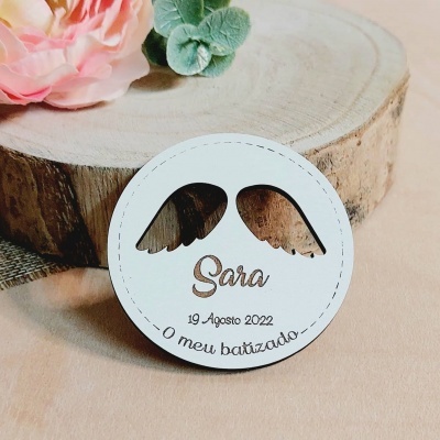 Medalha branca redonda personalizada com asas pretas e texto em português sobre madeira e flores