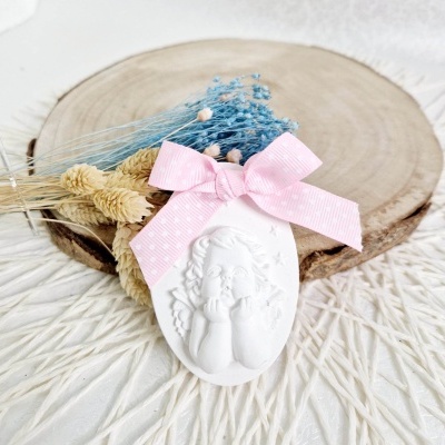 Ornamento branco em resina com anjo em relevo, laço rosa e raminhos secos sobre madeira e tecido branco