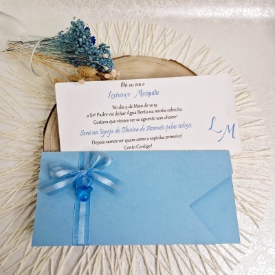 Convite branco com texto azul, envelope azul com laço, sobre prato de madeira e fundo texturizado
