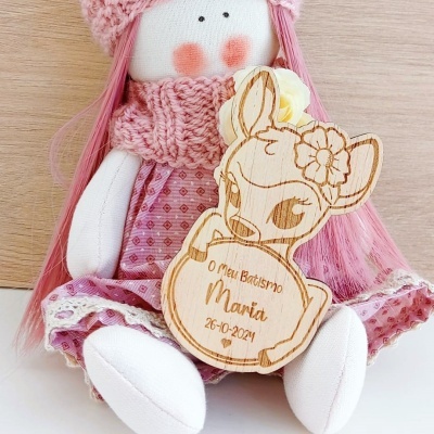 Boneca de tecido rosa segurando placa de madeira com gravura de veado e texto para batismo