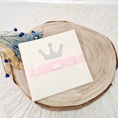 Convite branco com coroa prateada e fita rosa sobre base de madeira com flores secas