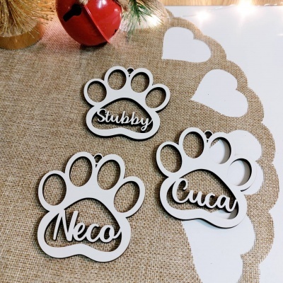 Ornamentos em forma de pata de animal com nomes Stubby, Neco e Cuca, sobre tecido de juta e superfície branca com corações recortados.