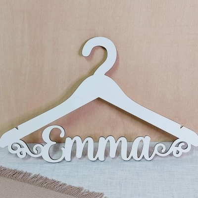 Cabide branco em madeira com nome 'Emma', sobre superfície clara e fundo de madeira