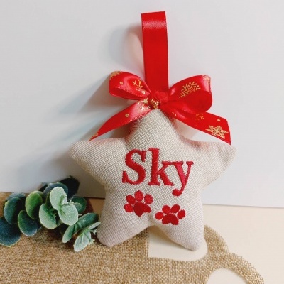 Enfeite de natal estrela bege com texto 'Sky' em vermelho e fita vermelha com flocos de neve dourados