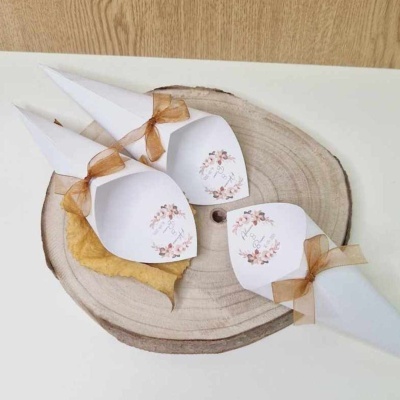 Porta-talheres em papel branco com fita castanha e etiquetas florais, sobre tábua de madeira clara com folha seca.